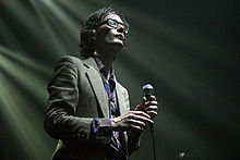 Jarvis Cocker—a Caucasian man with brown hair wearing glasses and a suit—holds a microphone onstage with his eyes closed.