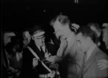 Waist high portrait of male in his forties, quite poor quality, taken before sunrise, wearing a light colored suit. Man in uniform to his right, onlooker at right.