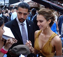 Smiling young woman with wavy hair pulled up in a loose bun, wearing a gold low-cut dress and accompanied by a man. She is signing autographs.
