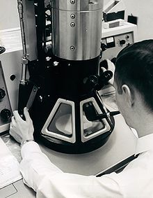 A black and white photograph of a young man examining a large microscope. The man has short, dark hair, is wearing a white shirt and a white lab coat and is holding a smoking pipe in his mouth. The microscope has a black conical base with three trapezoidal windows and a silver cylindrical body.