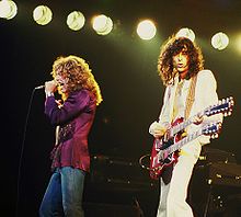 A colour photograph of Robert Plant with microphone and Jimmy Page with a double necked guitar performing onstage.