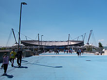 A straight tarmac road. At the head of the road is a stadium with a bowl-shaped outline, surrounded by a number of masts, with cables running to the stadium roof.