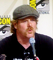 A bearded man wearing a black shirt and a gray beret sits in front a microphone. Sunglasses are han