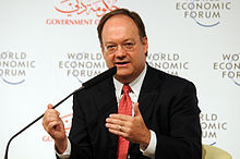 A bald middle aged man in a suit at a table speaks into microphone. Behind him several signs read World Economic Forum.