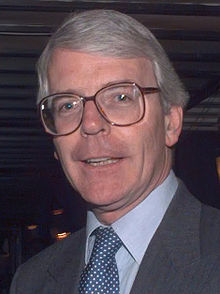 Head and shoulders of man in suit with grey hair in side parting, wearing large glasses with brown frame.