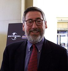 Headshot of a middle aged. Looking and smiling directly into the camera, the man wears rounded spectacles and sports a light grey beard. He wears a suit jacket, with a blue shirt and a patterned tie.