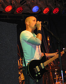 On stage, a man is holding a microphone to his mouth with his right arm while his left hand clasps the neck of a guitar. He is mostly bald headed, staring forward, with five coloured lights above and band equipment obscured behind.