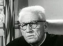 Black and white image of an older man with white hair and wearing glasses looking off to the right.