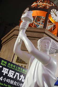 close-up view from below of a large white holding a torch; in upper background is large dome with clocks, a sign by the Hong Kong Federation of Students forms the lower part of the background
