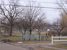 an image of a park. Many large trees are around the park and at the entrance a sign say Junge Park. Two basketball hoops and a baseball diamond are visible.