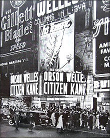 Night time view of a busy city street. Crowds gather on the sidewalk outside a cinema. A black limousine is parked outside the cinema. The entrance to the cinema has a pair of neon signs which reads "Orson Welles Citizen Kane". The rest of building is covered in neon advertising signs.