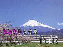 Over a background image of Mount Fuji and a Shinkansen train two lines of Japanese text. The first half of the first line is coloured red-violet, the rest white. The text reads "かみなりさまを　下 に聞く".