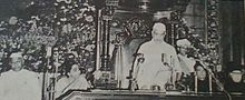 Photo of man in white cap/robe standing to speak at table