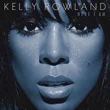 A close-up image of a woman's face covers the image. Her skin is blue and she is looking directly at the camera, with her face framed by her long black hair. Across the top in whit text it reads KELLY ROWLAND (the name of the artist). On the line immediately below, in smaller text and aligned to the right, it reads HERE I AM (the name of the album).