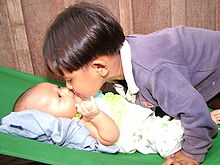 On the right a young boy of asiatic appearance with a pudding basin haircut, leans over a baby lying on its back on the left. The boy and baby are touching noses. The baby gazes up at the boy with an expression of intense interest.