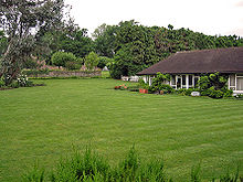 A white building with a black roof sits beside a large, well-kept lawn. Many trees are in the background of the picture, behind the building.