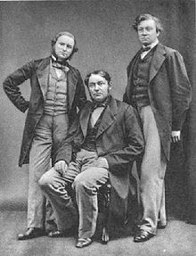  Three middle-aged men, with the one in the middle sitting down. All wear long jackets, and the shorter man on the left has a beard.
