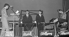 A black and white photograph of four members of Kraftwerk onstage, each with a synthesizer