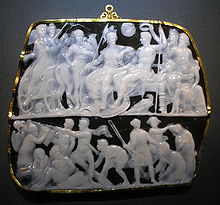 A photograph showing a roughly rectangular gem set in a gold frame with 2 carved panels with various figures carved in shallow from translucent white chalcedony against a solid black background