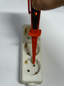 Screwdriver tester inserted in one pin of a European style electrical outlet, with an orange glow visible in the lamp.
