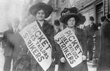 Two women in early 20th century clothing wear sashes that read, "Picket Ladies Tailer Strikers," while standing on a sidewalk in front of a building. A number of men stand on the sidewalk around them, some looking at the strikers, some facing away.