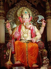 A sitting elephant-headed four-armed man statue, wearing gold ornaments, flower garlands and a orange dhoti.