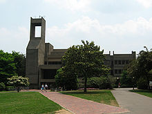 A large brutalist building with a tall boxlike structure at its front entrance siting in front of a wide green lawn with several trees and a brick walkway.