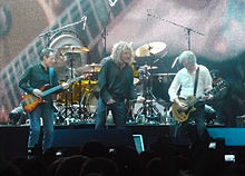 A colour photograph of John Paul Jones, Robert Plant and Jimmy Page performing onstage, with Jason Bonham partially visible on drums in the background