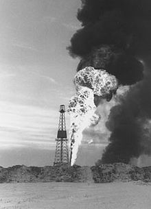 Older Black and white photo of an oil rig with a large flame and plume of smoke behind it.