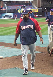 A man with a navy blue and red shirt with "Washington" written across the chest in white letters, gray pants, and a navy blue cap walking on a baseball field