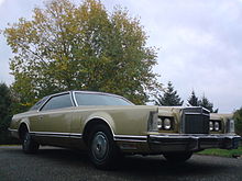 Front quarter view of a coupé parked in an open outdoor setting. The automobile is a sand Lincoln Continental Mark V.