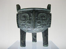 A heavily tarnished bronze bowl adorned with several carvings of squares that curl in on themselves at the bottom. It has three stubby, unadorned legs and two small, square handles coming off from the top rim.