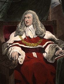 A middle age man sits in judicial robes, with a judge's wig on his head