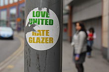 A photograph of a portion of a lamppost with a torn sticker that reads "Love United" in green above "Hate Glazer" in yellow.