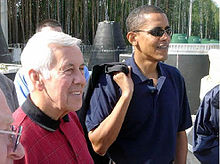 Gray-haired man and Obama stand, wearing casual polo shirts. Obama wears sunglasses and holds something slung over his right sholder.