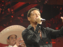 A man sings into a microphone. He is wearing a black shirt. Behind him, a man wearing a white sombrero plays on a silver trumpet.