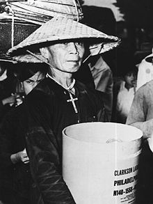 Wrinkled old man wearing a straw conical hat, black ao baba, and a large crucifix necklace holds a white cylindrical container with English language writing on it. Behind him is another person, partially obscured, wearing black with straw baskets of luggage on their head.