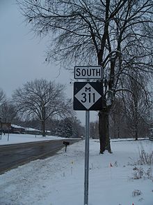 A highway sign along side the road on a snowy day.