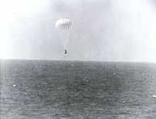 A conical black spacecraft falling towards the surface of the ocean under a single white parachute, seen from some distance away. Very little detail can be seen.