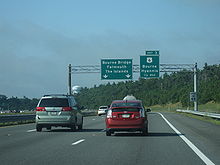 Ground-level view of three lanes of a divided expressway; two large green exit signs are visible in the distance, and the road is surrounded by dense forests.