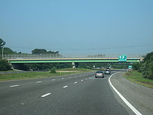 Ground-level view of a three lanes of a divided freeway; a large green and gray overpass bridge and a green exit sign are visible in the distance.