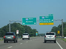 Ground-level view of three lanes of a divided freeway with two green exit signs directly overhead; the sign on the right is marked with a bright yellow banner that reads "Exit Only".