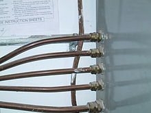 Five metal-covered cables enter the left side of an electrical panel through brass fittings, with bare cable running down the concrete wall behind.