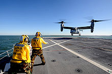  A V-22 performing a vertical landing on the USS New York with two of the ship's crew nearby.