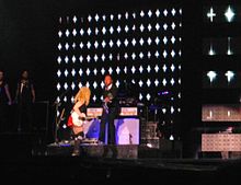 A faraway image of a blond woman, wearing a red shorts, black T-shirt and playing a white electric guitar. Her face is away from the image. Beside her, a band stands with the backdrops displaying white stars on black.