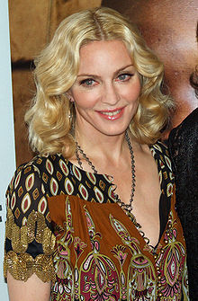 Upper body of a middle-aged blonde woman. Her hair is parted in the middle and falls in waves to her shoulder. She is wearing a loose dress with black and brown prints on it. A locket is hung around her neck, coming up to her breasts. She is looking to the right and is smiling.
