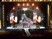 Distant shot of a concert stage. An arch with spotlights stands above a life-size locomotive and two big screens which display the male singer in left profile. Below and in front of the locomotive is musical equipment and four band members. At extreme left is a male playing guitar he wears a school boy's uniform. Central stage has two guitarists with a drummer and his kit between them. Front stage has a catwalk with the singer turned to his left profile, he is cradling the microphone in his right hand and has his left fist near his face. He wears a cap, dark singlet and jeans.