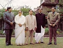 Group portrait of four men and one woman standing in a garden, on a shortly-cropped lawn. Two of the men and the woman are wearing Indian-style dress; the man in the centre and the one on the far right are wearing Western-style business suits.