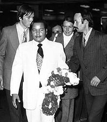 A self-confident young man wearing a white suit, white shirt and dark tie, his left arm holding Indian-style flower garlands; several young Westerners in business suits are standing behind him.