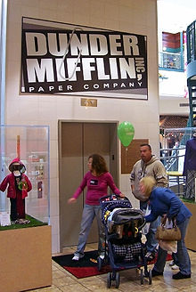 Three adults and a child in a stroller in front of a set of closed elevator doors. Above them is a sign with "Dunder Mifflin Paper Company" on it in white letters on a black background